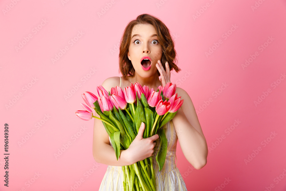 portrait of surprised woman with bouquet of spring tulips looking at camera isolated on pink