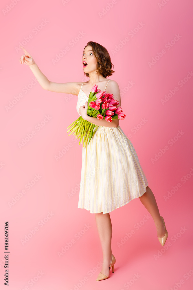 shocked woman with bouquet of pink tulips pointing away isolated on pink