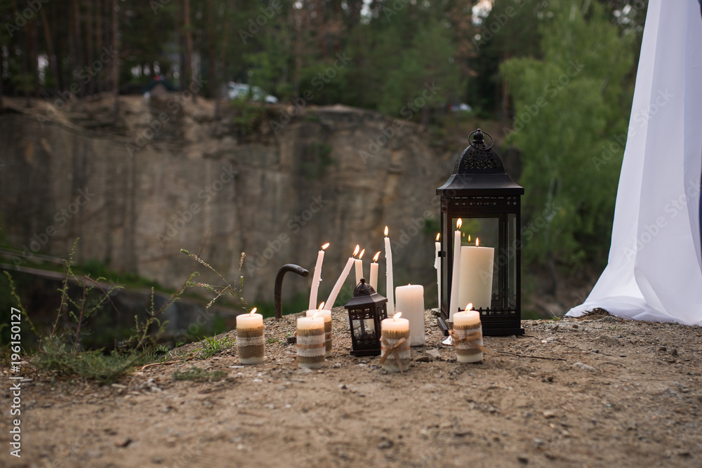 A lot of white burning candles on candlesticks and in lanterns in forest near the wedding arch with white material A lot of white burning candles on candlesticks and in lanterns in forest near the wedding arch with white material