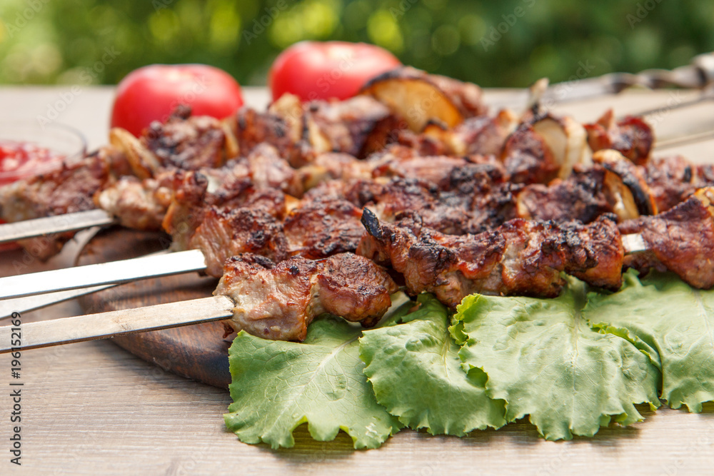 Grilled pork on skewers roasted on the grill and lettuce leaves on wooden chopping board with tomato sauce in a bowl and tomatoes on the background