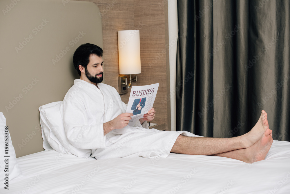 smiling businessman in bathrobe reading newspaper while relaxing on bed at hotel suite smiling businessman in bathrobe reading newspaper while relaxing on bed at hotel suite