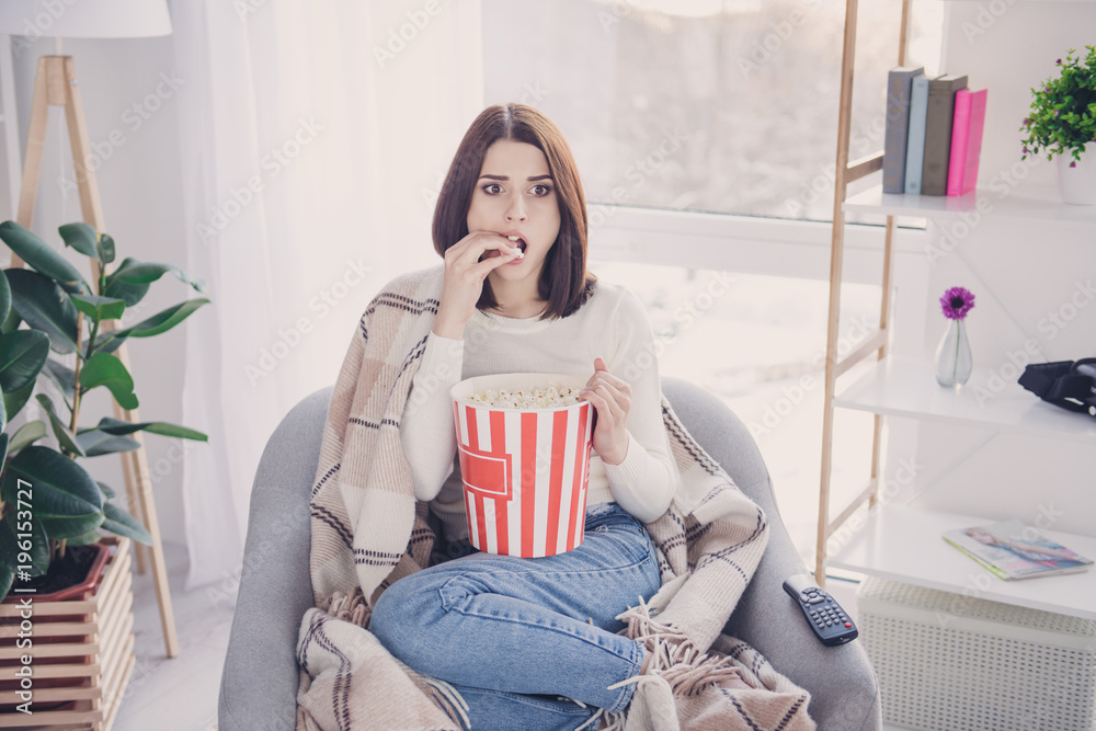 Naklejka premium Portrait of pretty, charming, stylish, exciting girl with modern hairstyle putting pop corn in her open mouth watching spectacular film, covered with plaid, wearing denim outfit, jeans and pullover