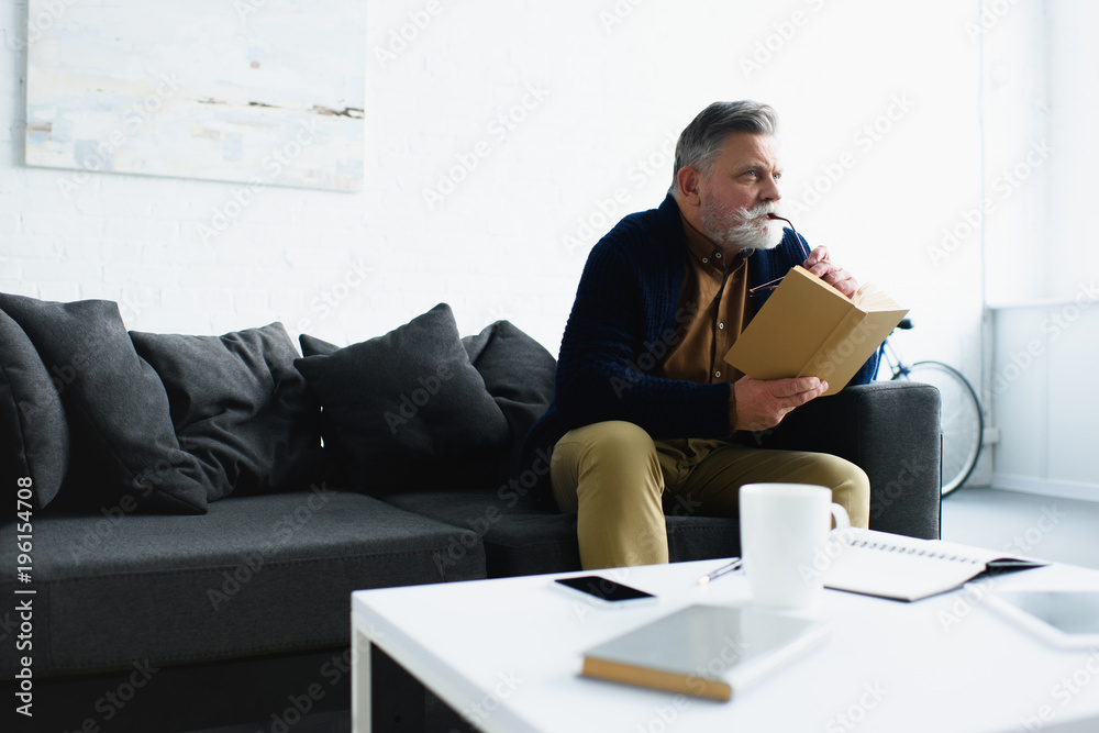 pensive bearded senior man holding book and looking away at home pensive bearded senior man holding book and looking away at home