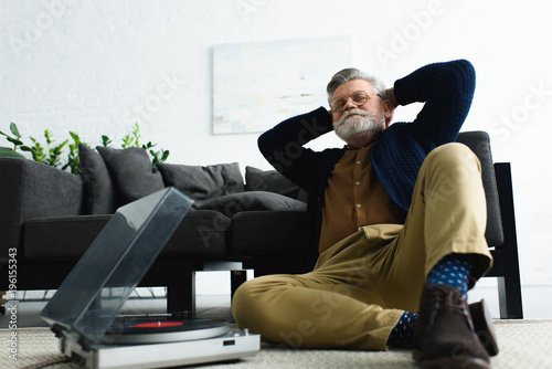 relaxed stylish man in eyeglasses sitting with hands behind head and listening music with vinyl record player at home