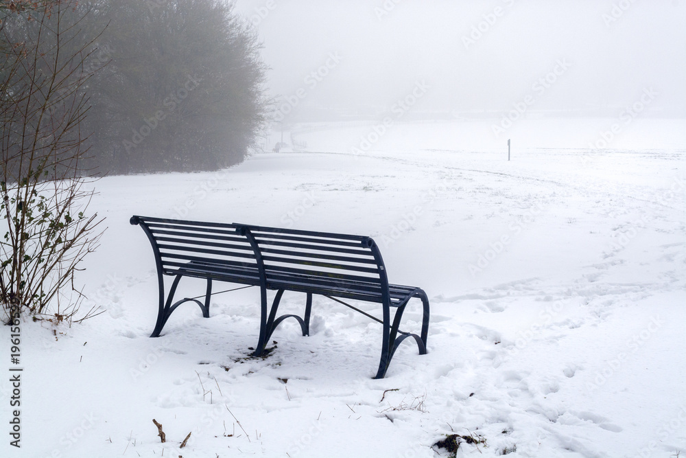 A bench at the edge of snow-covered field on foggy morning - winter scenery