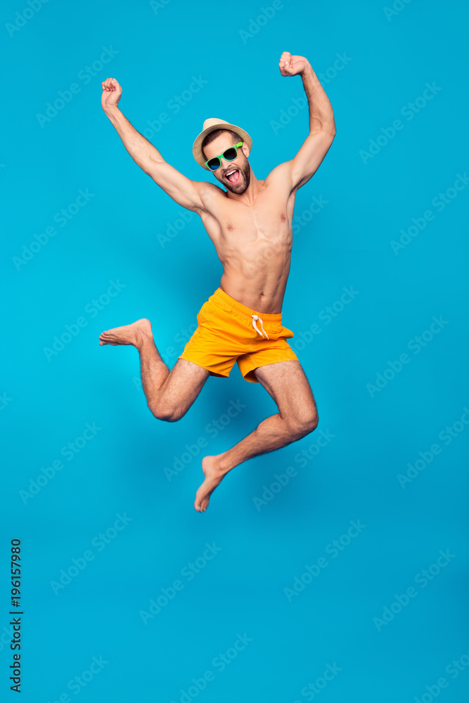 Fullbody, full length portrait of attractive, successful, cheerful, foolish, comic ladies' man in yellow shorts, jumping with open mouth and raised hands, isolated on blue background