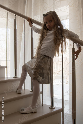 Charming young girl with long hair in smart dress posing on stairs in house. Expressive cute face close-up and funny grimaces. Age of innocence. The concept of happy childhood  home leisure  friends