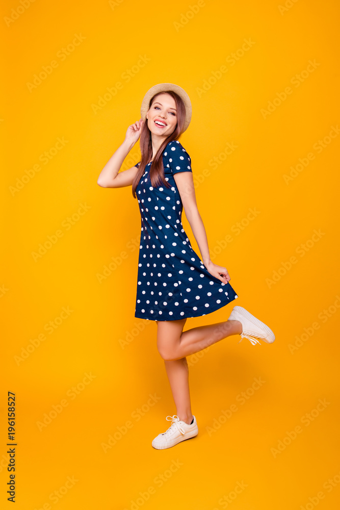 Full-length, fullbody portrait of nice, cheerful, positive, cute girl with raised leg, holding hands on hat and bottom of polka-dot dress, posing over yellow background Full-length, fullbody portrait of nice, cheerful, positive, cute girl with raised leg, holding hands on hat and bottom of polka-dot dress, posing over yellow background