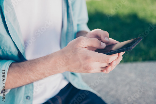 Modern technology people remote work job video concept. Cropped close up photo of handsome man's hands typing message on new stylish smartphone wearing jeans denim casual shirt green grass