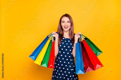 Portrait of glad, charming, pretty, crazy girl happy, excited that bought a lot of clothes at low prices, holding packets in two raised hands, isolated on yellow background, unbelievable shopping