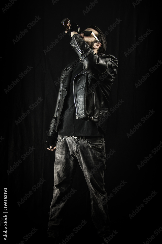 A man wearing a black leather jacket and sunglasses, drinking from a bottle of beer. A man wearing a black leather jacket and sunglasses, drinking from a bottle of beer.