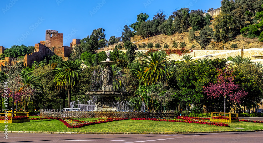 Obraz premium Fountain (Fuente de las tres gracias) at the end of Paseo del Parque with the castle (alcazaba) to rear. Malaga city, Spain.
