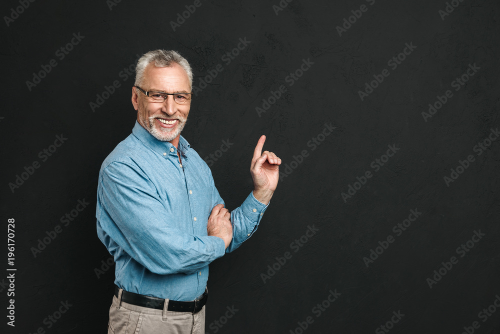Portrait of caucasian retired man 60s with grey hair and beard in shirt smiling and pointing index finger aside on copyspace, isolated over black background Portrait of caucasian retired man 60s with grey hair and beard in shirt smiling and pointing index finger aside on copyspace, isolated over black background