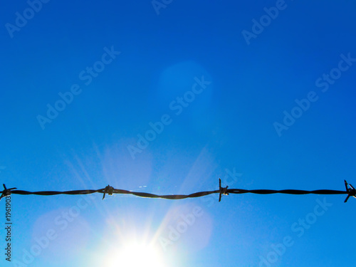 Wire fencing against a blue sky and sun. Black horizontal line of sharp-cut silhouette and bright sunlight.