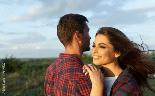 Horizontal shot of happy couple have fun on a nature background. Rear view of handsome Caucasian guy wearing plaid shirt with smiling girlfriend hugs him. Lifestyle, people and travel concept.