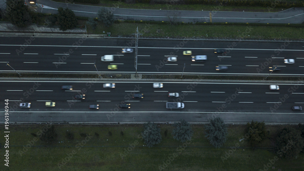 Aerial night view of a highway junction in Rome, Italy. The street crosses an industrial area. On the road, many cars drive at high speed with the headlights on. Around there are grass and trees.