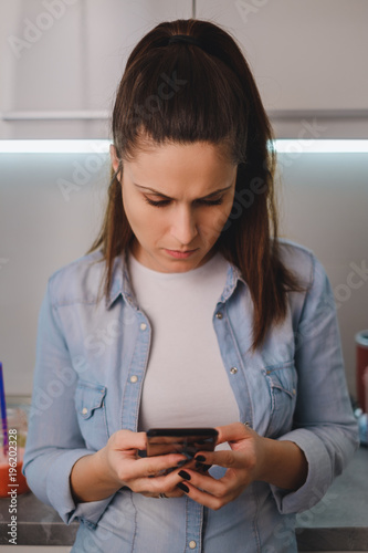 фотографія Angry girl looking at mobile phone in the kitchen