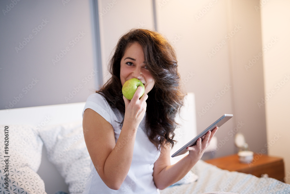 Fototapeta premium Close up of smiling beautiful middle aged woman sitting on the bed with a tablet and bites an apple while looking at the camera.