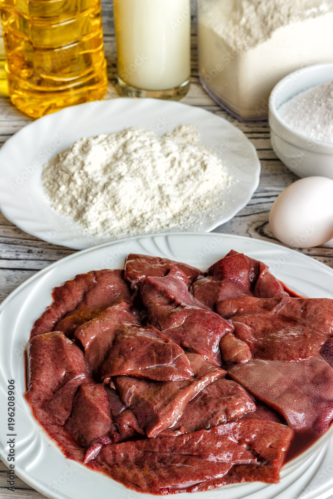 Fototapeta premium Raw liver on a white plate on a wooden background. A set of products for cooking fried liver egg oil salt flour. Food ingredient. Selective focus.
