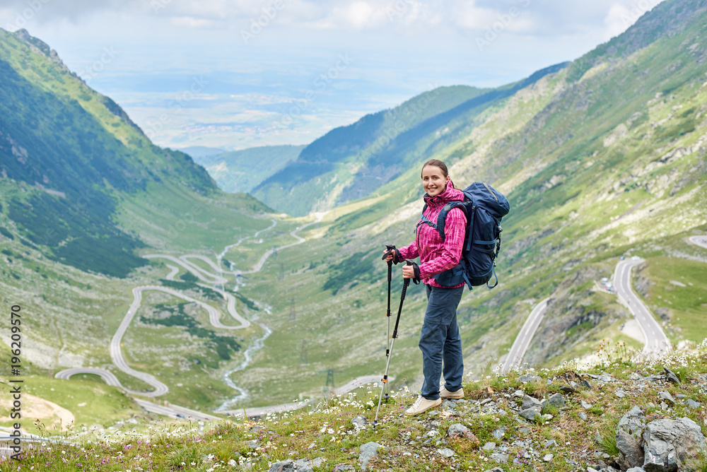 Smiling female tourist standing with walking stick to beautiful view of magnificent winding Transfagarashan road in Romania. Woman climber green hills mountains happy rock travel route path