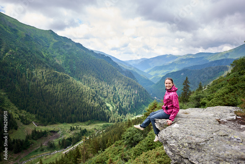 Happy beautiful woman tourist sitting on rock edge, looking to the camera, enjoying breathtaking view of green grassy slopes and mountains with trees, fir trees and pines in Romania