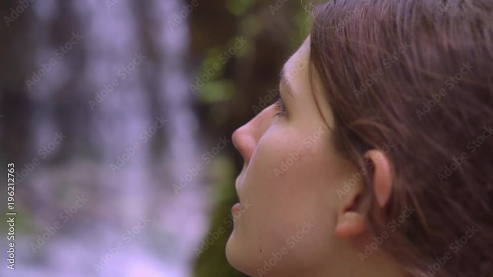 Young girl looks up at waterfall in background of forest.
Young girl looks up at waterfall in background of forest.