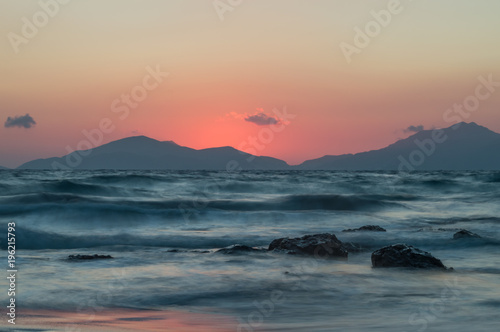 A long exposure of the sea at golden hour, as the sun sets behind the mountains. The sea looks ethereal as it breaks over the rocks and beach.