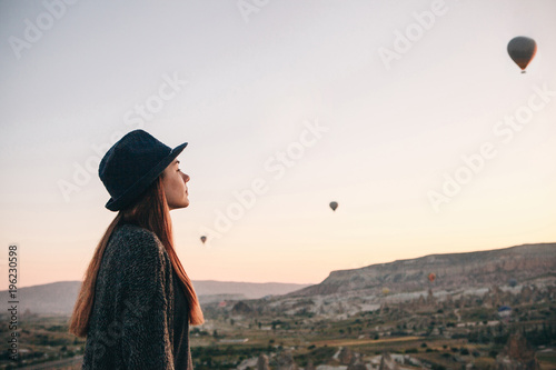A tourist girl in a hat admires hot air balloons flying in the sky over Cappadocia in Turkey. Impressive sight.