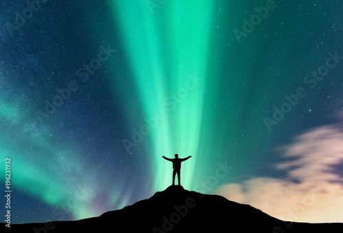 Aurora and silhouette of standing man with raised up arms on the mountain. Lofoten islands, Norway. Aurora borealis and happy man. Sky with stars and green polar lights. Night landscape with aurora