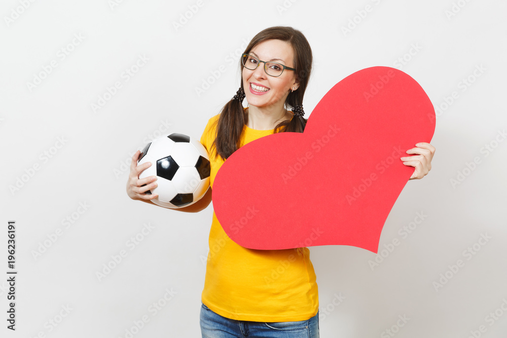 Smiling European woman, fun pony tails, football fan or player in glasses,  yellow uniform hold classic soccer ball, red heart isolated on white  background. Sport, football, healthy lifestyle concept. Stock Photo
