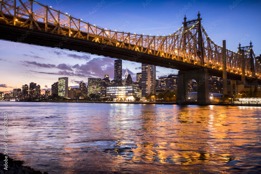 The lights of New York City and the Queensboro Bridge reflect off the water of the East River as the sun sets over the city.