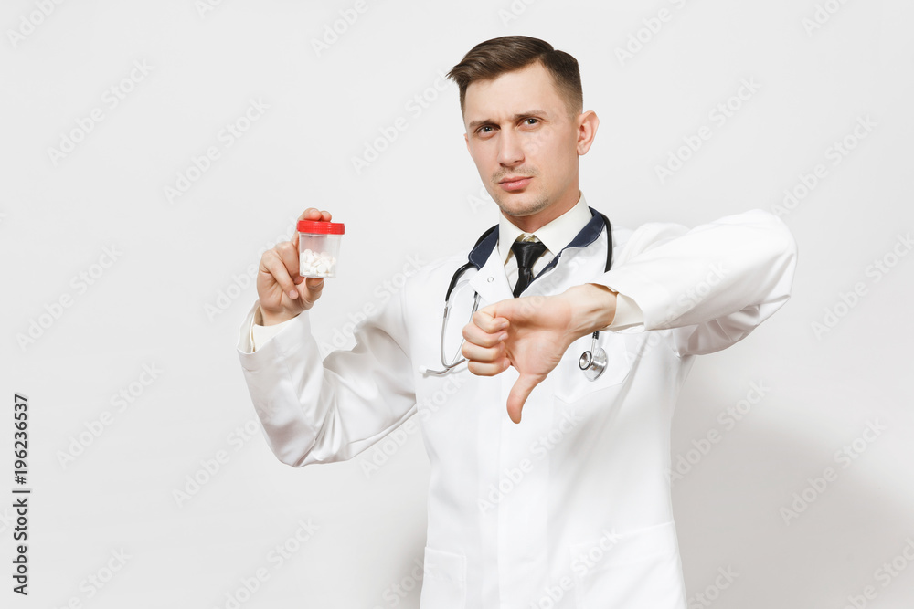 Serious young doctor man showing thumbs down isolated on white background. Male doctor in medical uniform, stethoscope holding bottle with white pills. Healthcare personnel, health, medicine concept. Serious young doctor man showing thumbs down isolated on white background. Male doctor in medical uniform, stethoscope holding bottle with white pills. Healthcare personnel, health, medicine concept.