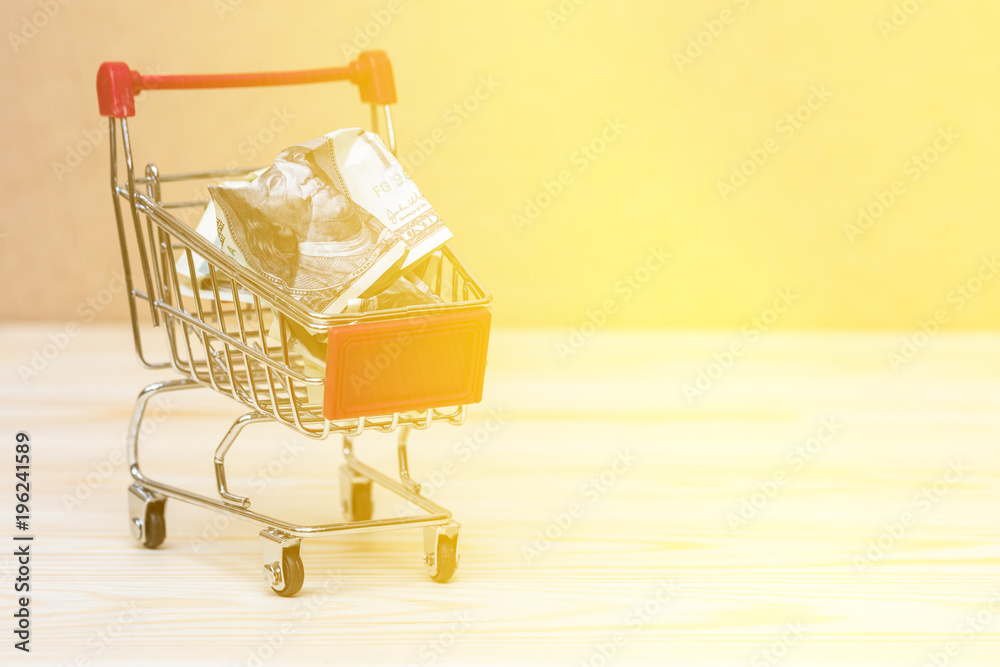Shopping cart on wooden surface with crumpled up dollar bill. Symbol of consumerism with lighting effects