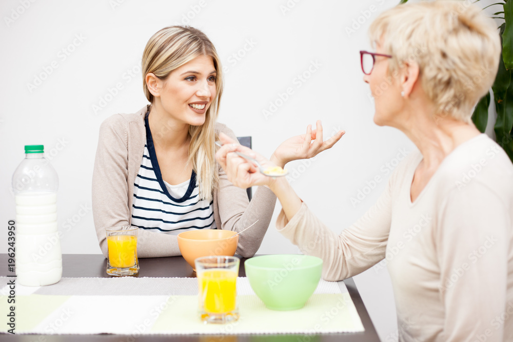 Senior mother and daughter talking and eating healthy cereal breakfast in brightly lit dining room, Focus on daughter. Happy family moments at home Senior mother and daughter talking and eating healthy cereal breakfast in brightly lit dining room, Focus on daughter. Happy family moments at home
