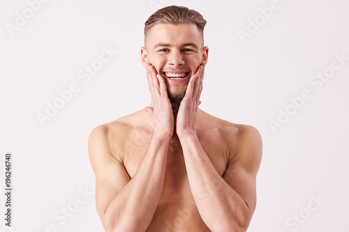 Portrait of young naked man applying aftershave and smiling while standing isolated on white background. Bearded hispter man standing shirtless and touching his face with hands. Men's skin care photo