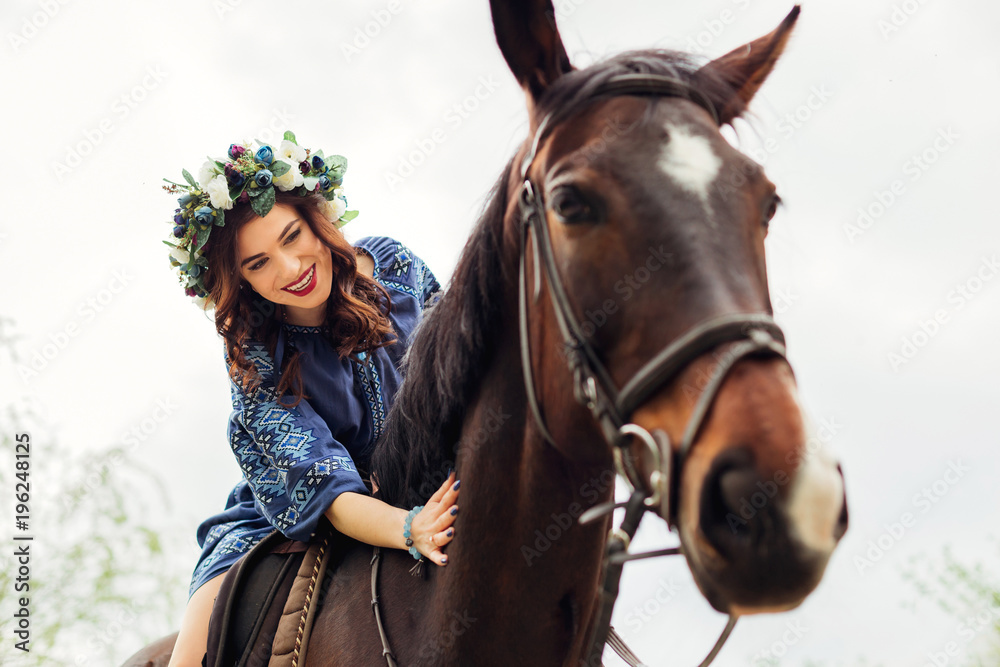 Fototapeta premium close-up of a cute girl with a wreath on her head sitting on a horse and smiling and stroking a horse's neck