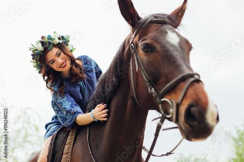 cute girl with a wreath on her head and a beautiful hairstyle that sits on a horse and caress a horse's neck
