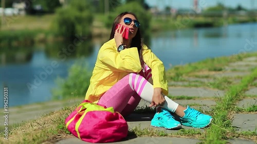 Sportswoman in yellow sports jacket sits near pond and talks on cell phone. Girl in sportswear sits near lake and speaks on smartphone photo