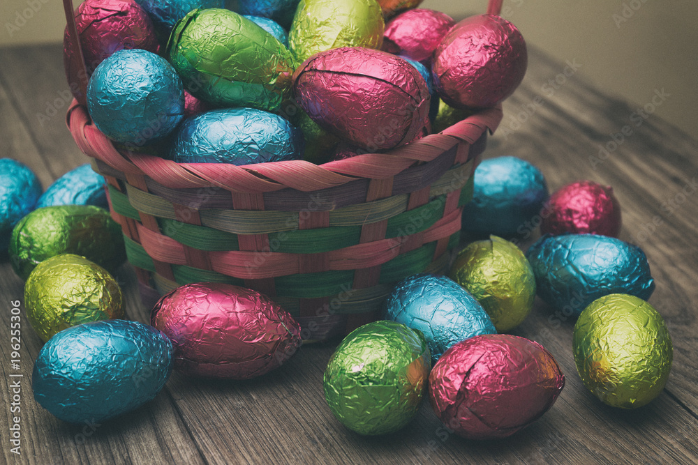 Straw basket filled with Easter chocolate eggs wrapped in colorful tinfoil on top of a wooden table Straw basket filled with Easter chocolate eggs wrapped in colorful tinfoil on top of a wooden table