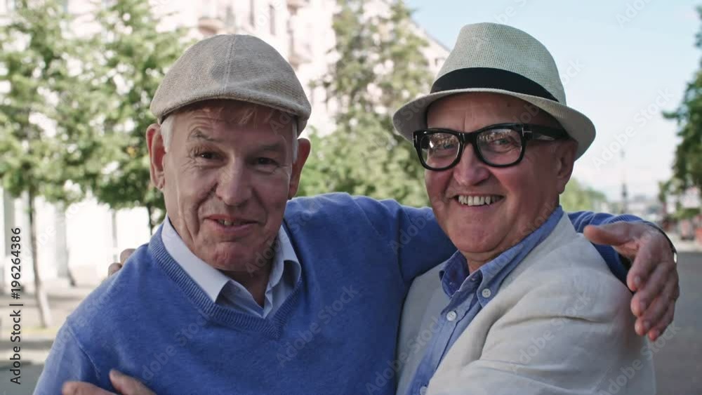 Medium shot of two senior men in hats standing in pedestrian street on warm day, hugging each other and patting on the shoulders Medium shot of two senior men in hats standing in pedestrian street on warm day, hugging each other and patting on the shoulders