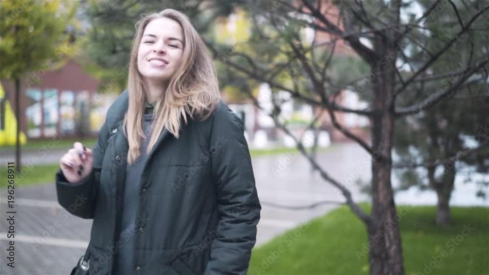 Happy young woman with fair hair wearing a coat and holding a bag is dancing in an autumn street. Handheld slow motion medium shot
