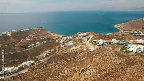 Flying down the hill with luxury white villas, swimming pools surrounded by brown stones, boulders, arid bushes, dusty roads with cars towards Aegean sea coastline. Cyclades. Faros Armenistis, Mykonos