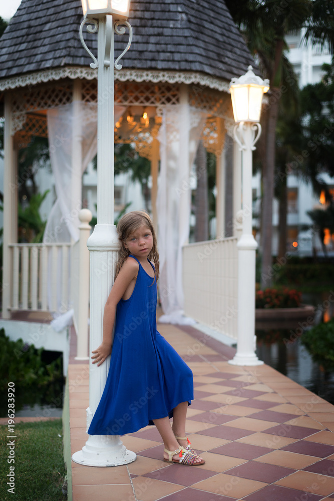 Cute blonde Caucasian little girl in blue dress leans against a pillar . Evening scene with gazebo is on the background. Cute blonde Caucasian little girl in blue dress leans against a pillar . Evening scene with gazebo is on the background.