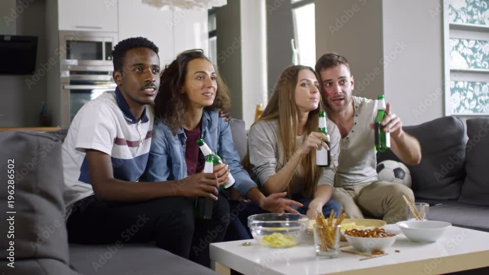 Two young mixed-raced couples sitting together on couch in the living room, watching soccer match on TV, drinking beer and laughing Two young mixed-raced couples sitting together on couch in the living room, watching soccer match on TV, drinking beer and laughing