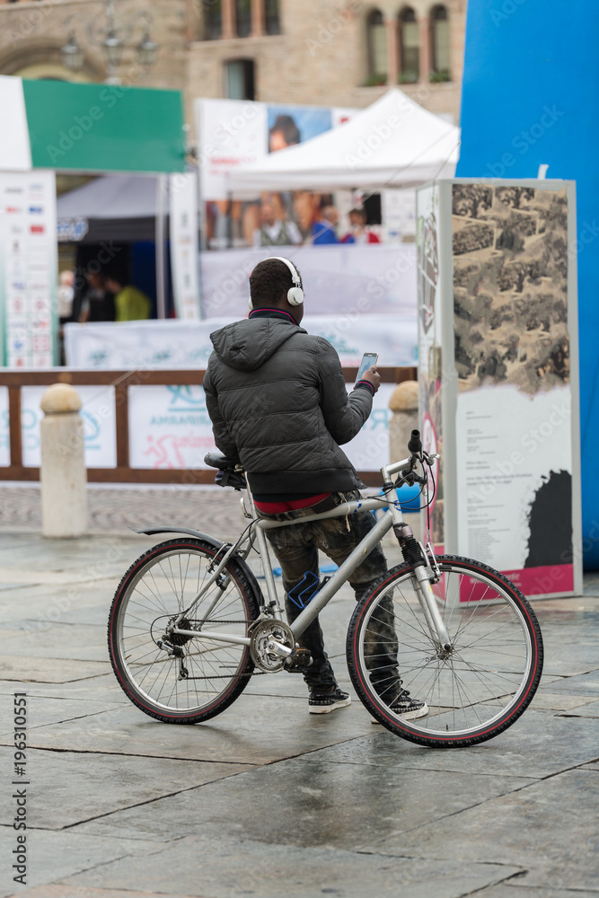 Rear View of  Young Man with EarPhones Sitting on Bicycle and Using Smartphone on City Street: People, Style, Technology, Leisure and Lifestyle