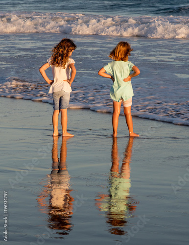 Two little girls playing in the shallow water of the sea late in the afternoon. Their reflections are clearly visible on the wet sand. The wind is blowing through their long hair
