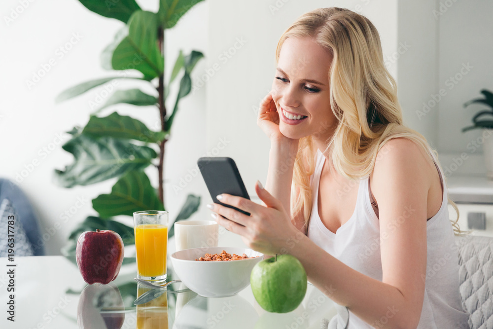beautiful smiling blonde girl using smartphone while having breakfast at home beautiful smiling blonde girl using smartphone while having breakfast at home