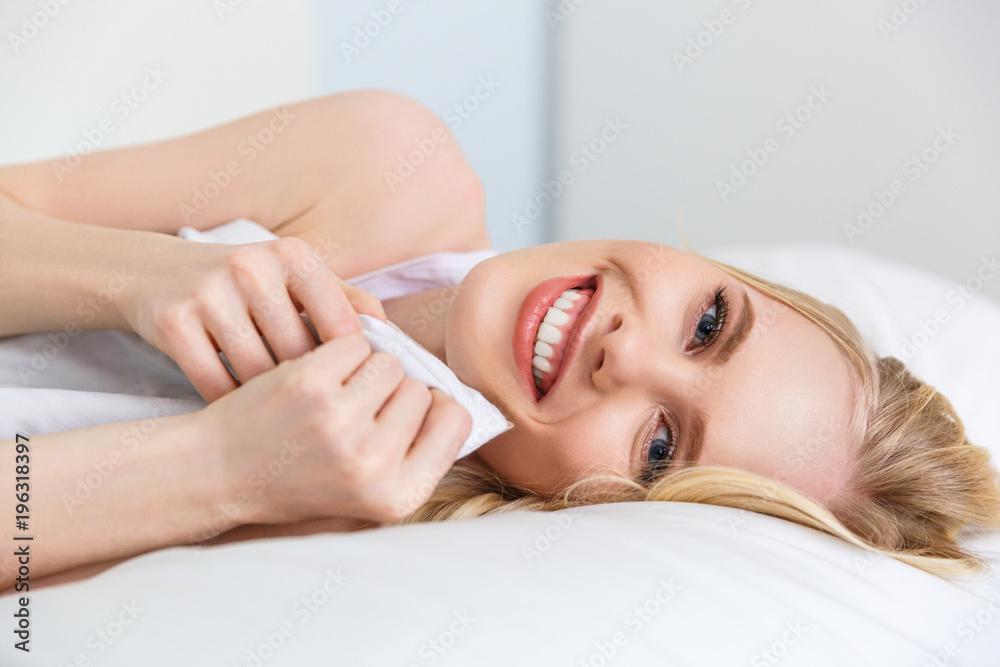 attractive blonde girl holding blanket and smiling at camera while lying on bed attractive blonde girl holding blanket and smiling at camera while lying on bed
