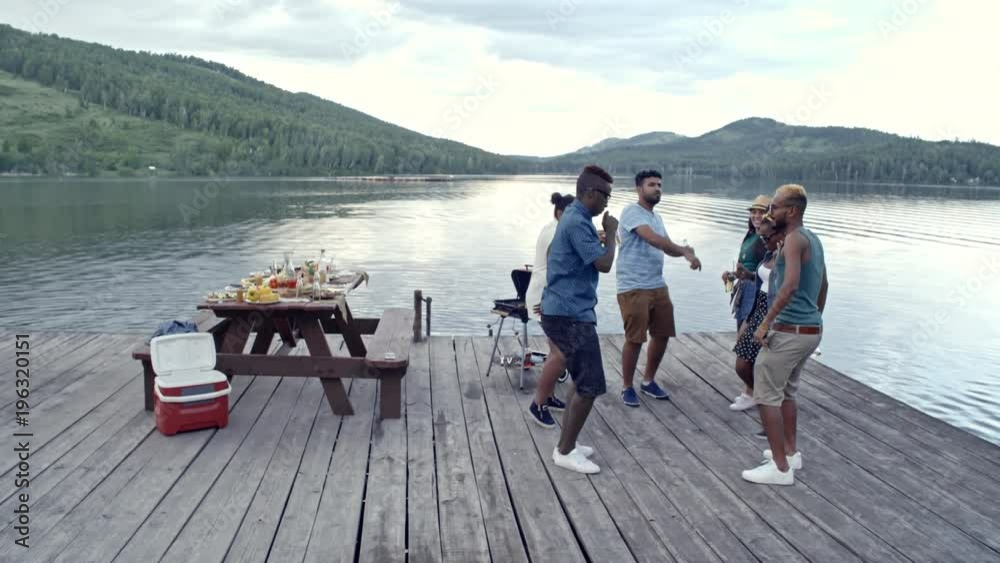 Tilt down shot of six young friends having picnic and party outdoors and dancing on wooden dock by lake Tilt down shot of six young friends having picnic and party outdoors and dancing on wooden dock by lake