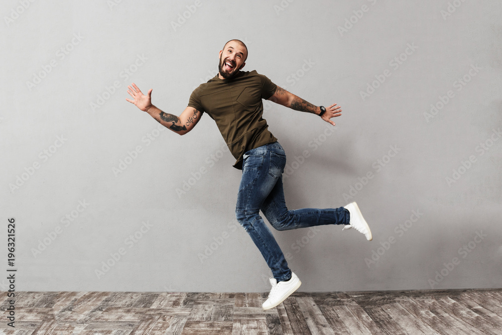 Full-length image of smiling guy with tattoo on his arms having fun while posing on camera, isolated over gray background
